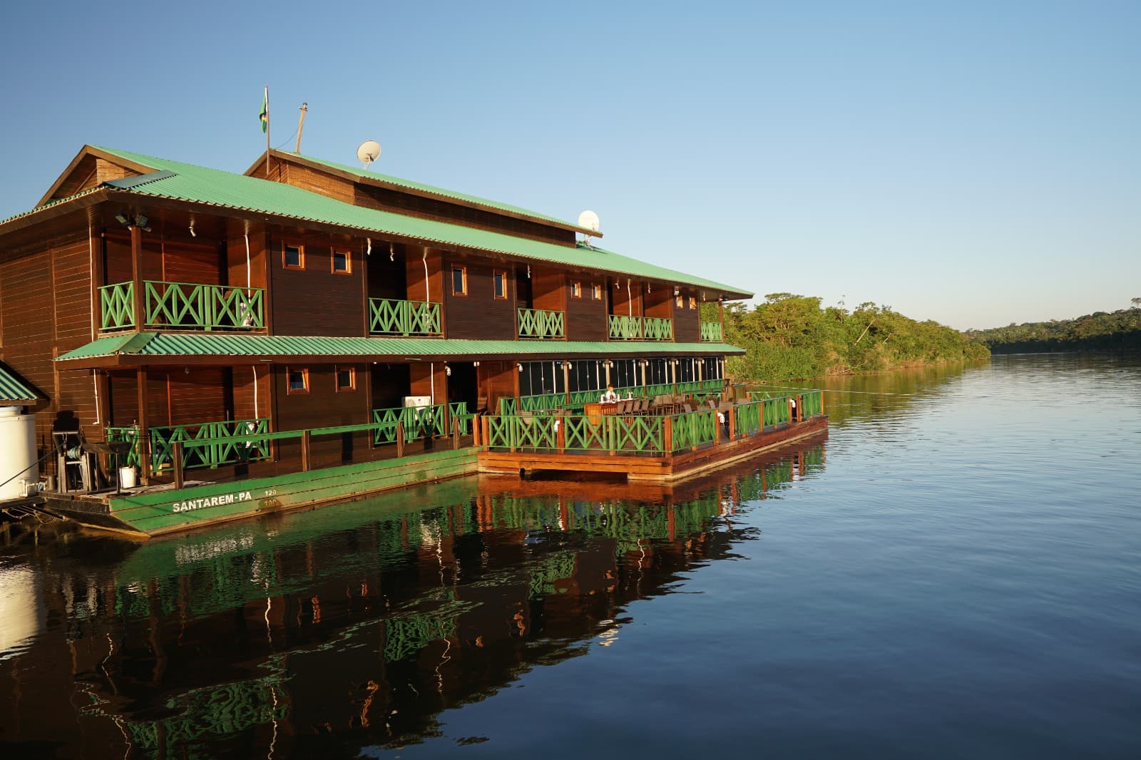 Fachada do Ecolodge da Barra ao entardecer — flutuante de dois andares em madeira com telhado verde refletido no Rio Tapajós